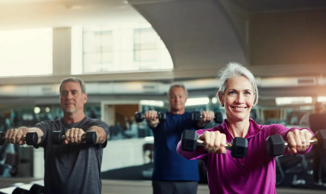 Three older adults, a smiling white-haired woman in a magenta top and two men, doing standing dumbbell raises together in a bright gym.